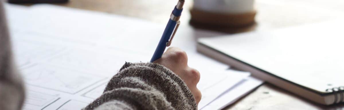 person writing on brown wooden table near white ceramic mug