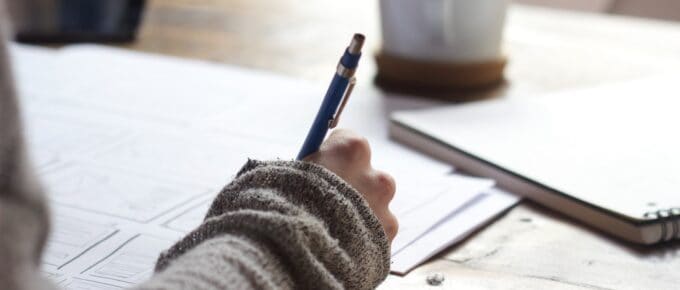 person writing on brown wooden table near white ceramic mug